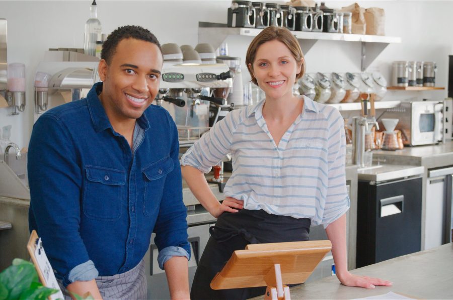 Portrait Of Smiling Male And Female Owners Or Workers Behind Counter In Coffee Shop